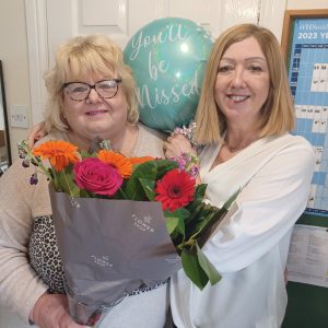 A photo of Glenda and Yvonne. Glenda on the left is holding flowers. Yvonne is holding a blue balloon that says "you'll be missed"