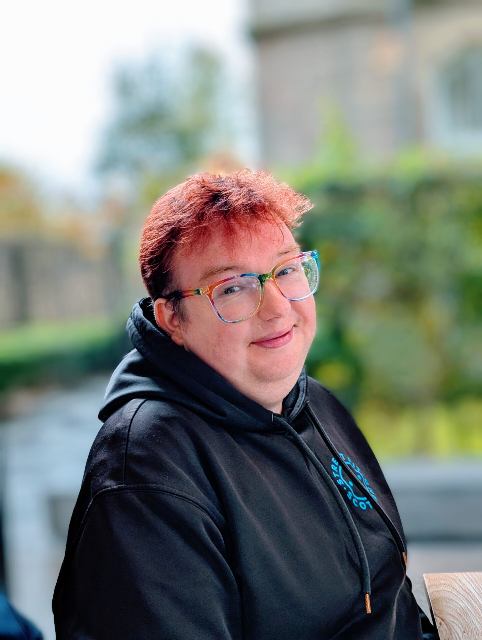 Joseph is smiling at the camera and wearing a black hoodie and rainbow coloured glasses with greenery in the background.