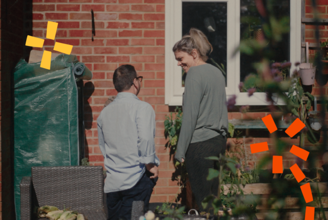 The image shows two people in a garden: on the left, a man wearing a shirt and glasses, his back is to the camera. On the right, a lady with blonde hair in a pony tail, she has her back to the camera but is smiling.